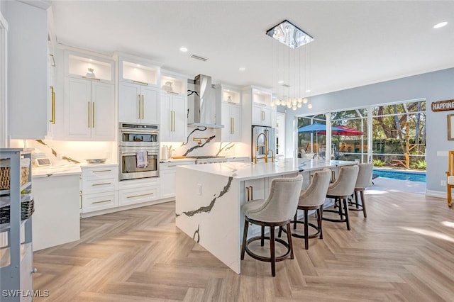 kitchen featuring a center island with sink, wall chimney range hood, decorative light fixtures, white cabinetry, and stainless steel appliances