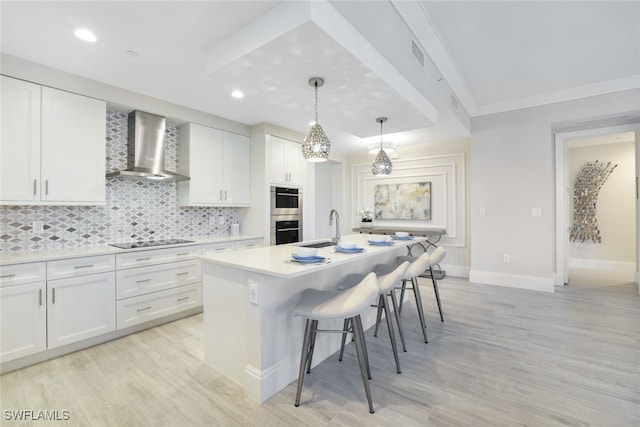 kitchen with white cabinetry, wall chimney range hood, a kitchen breakfast bar, a center island with sink, and light wood-type flooring