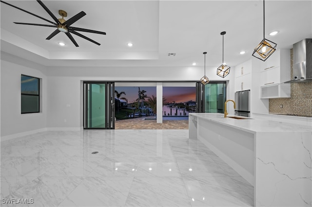 kitchen featuring wall chimney range hood, hanging light fixtures, backsplash, white cabinetry, and light stone countertops