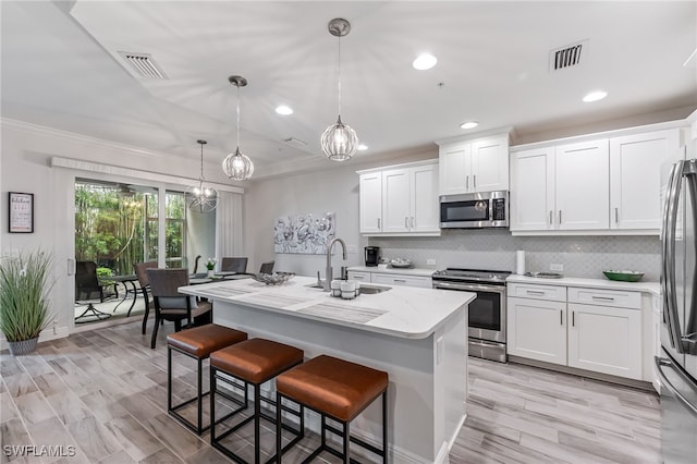 kitchen featuring stainless steel appliances, ornamental molding, hanging light fixtures, white cabinetry, and a center island with sink