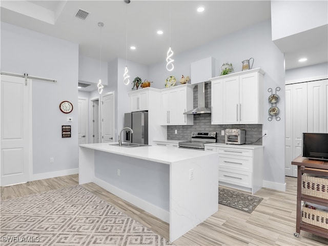 kitchen with wall chimney range hood, appliances with stainless steel finishes, a kitchen island with sink, a barn door, and pendant lighting