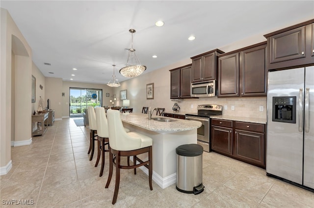 kitchen with dark brown cabinets, light stone countertops, hanging light fixtures, appliances with stainless steel finishes, and an island with sink