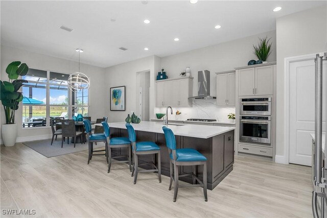 kitchen with wall chimney exhaust hood, a kitchen breakfast bar, light hardwood / wood-style flooring, double oven, and a center island with sink