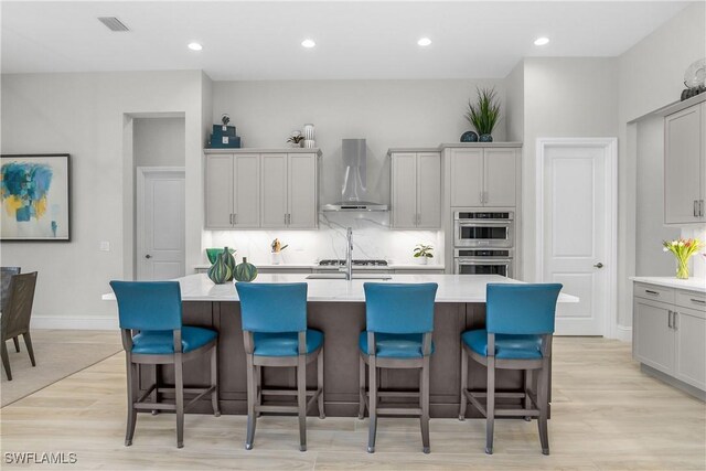 kitchen featuring a kitchen bar, a kitchen island with sink, wall chimney range hood, and light wood-type flooring