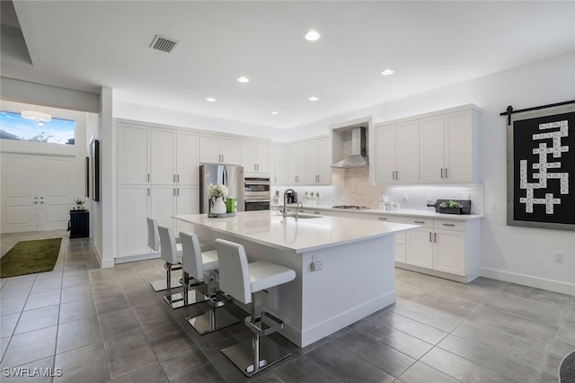 kitchen with a kitchen island with sink, wall chimney range hood, sink, a barn door, and appliances with stainless steel finishes