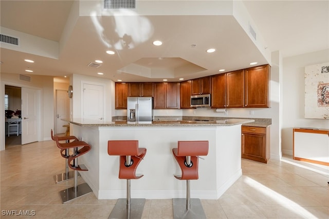 kitchen featuring a kitchen island with sink, a tray ceiling, a breakfast bar, light stone countertops, and appliances with stainless steel finishes