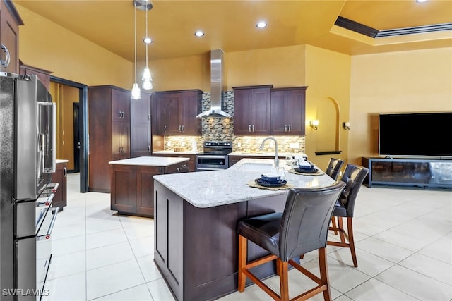 kitchen with decorative backsplash, wall chimney exhaust hood, an island with sink, pendant lighting, and appliances with stainless steel finishes