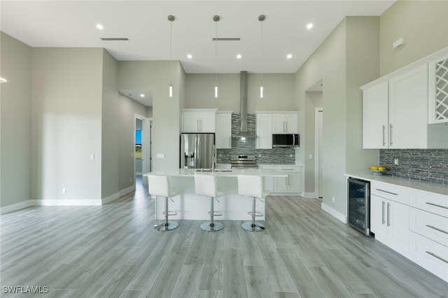 kitchen featuring a breakfast bar area, beverage cooler, a towering ceiling, appliances with stainless steel finishes, and wall chimney range hood