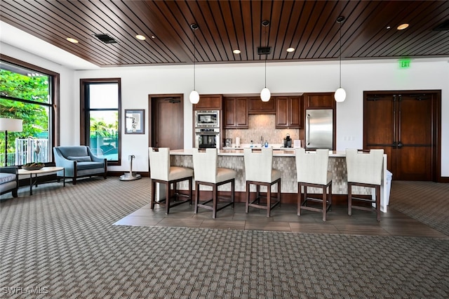 kitchen with a breakfast bar area, backsplash, hanging light fixtures, built in appliances, and wooden ceiling