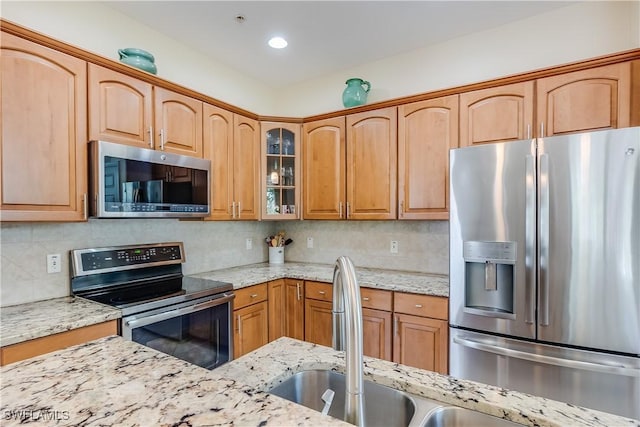 kitchen with glass insert cabinets, backsplash, light stone countertops, stainless steel appliances, and a sink