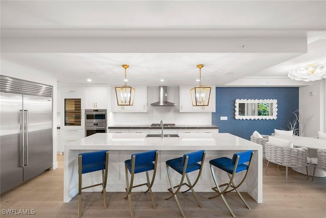 kitchen with light wood-style floors, wall chimney range hood, white cabinets, appliances with stainless steel finishes, and a notable chandelier