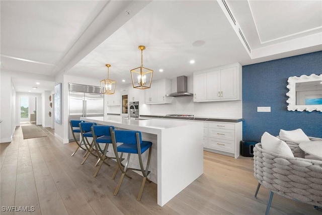 kitchen featuring wall chimney range hood, light wood-style flooring, cooktop, a kitchen bar, and built in refrigerator