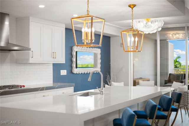 kitchen with sink, wall chimney range hood, white cabinetry, decorative light fixtures, and backsplash