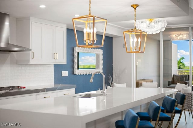 kitchen with wall chimney range hood, decorative backsplash, an inviting chandelier, a sink, and stainless steel gas stovetop