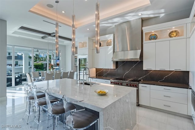 kitchen with an island with sink, tasteful backsplash, a raised ceiling, wall chimney exhaust hood, and high end stainless steel range oven