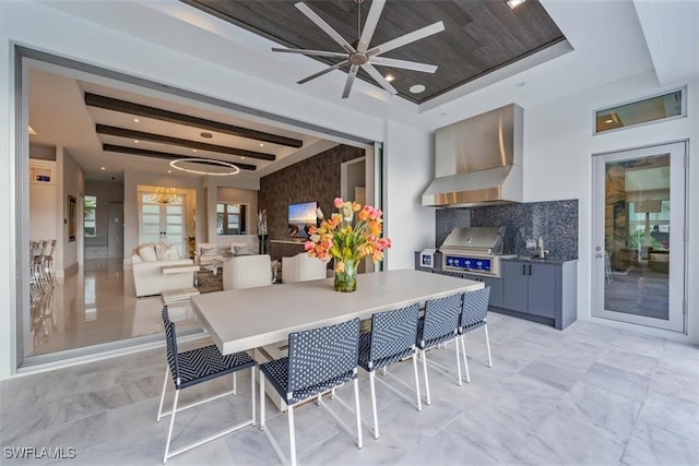 kitchen with backsplash, wall chimney range hood, a tray ceiling, ceiling fan with notable chandelier, and a kitchen bar