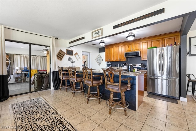 kitchen with light tile patterned flooring, a kitchen bar, ceiling fan, stainless steel appliances, and light stone counters