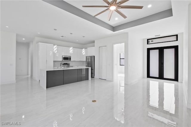 kitchen with ceiling fan, stainless steel appliances, a raised ceiling, a center island with sink, and white cabinets