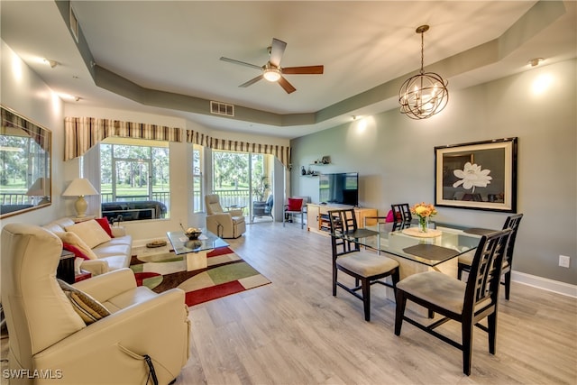 dining space with ceiling fan with notable chandelier, a tray ceiling, and light hardwood / wood-style floors