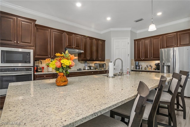 kitchen with a spacious island, ornamental molding, appliances with stainless steel finishes, decorative light fixtures, and light stone counters