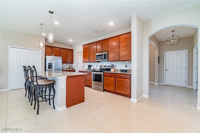 kitchen featuring a kitchen island with sink, light stone counters, decorative backsplash, hanging light fixtures, and appliances with stainless steel finishes