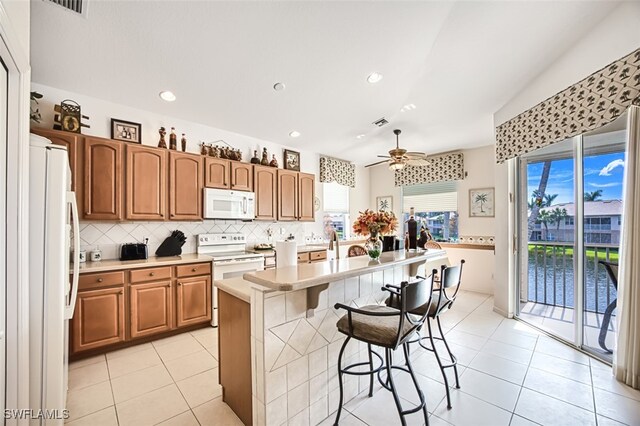 kitchen featuring a center island with sink, a breakfast bar, ceiling fan, white appliances, and tasteful backsplash