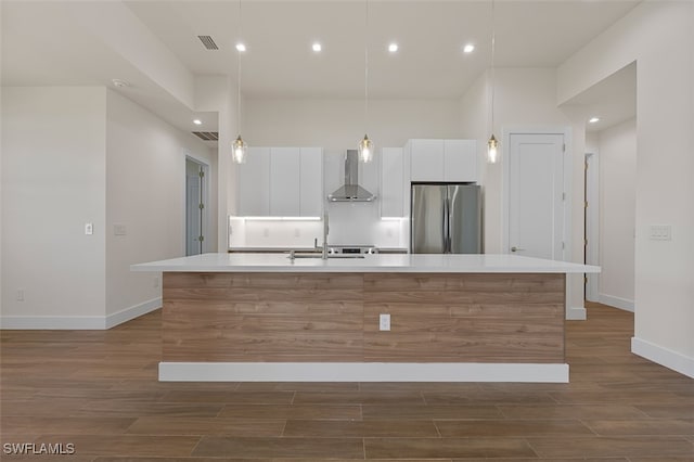 kitchen featuring stainless steel refrigerator, a kitchen island with sink, wall chimney exhaust hood, and white cabinets