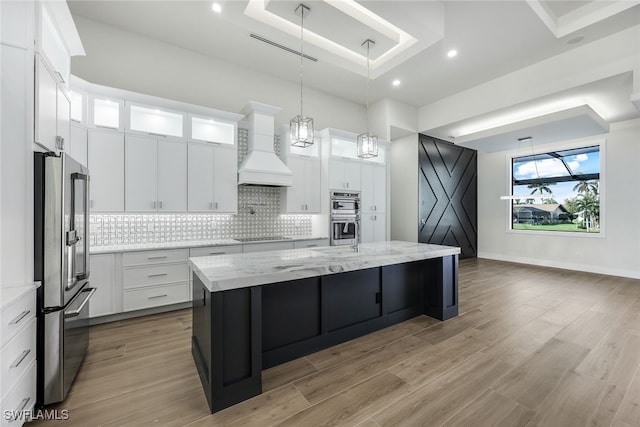 kitchen with glass insert cabinets, premium range hood, white cabinetry, appliances with stainless steel finishes, and a center island with sink