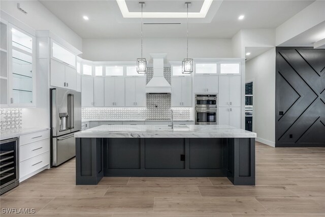 kitchen featuring custom exhaust hood, beverage cooler, a large island with sink, appliances with stainless steel finishes, and light stone counters