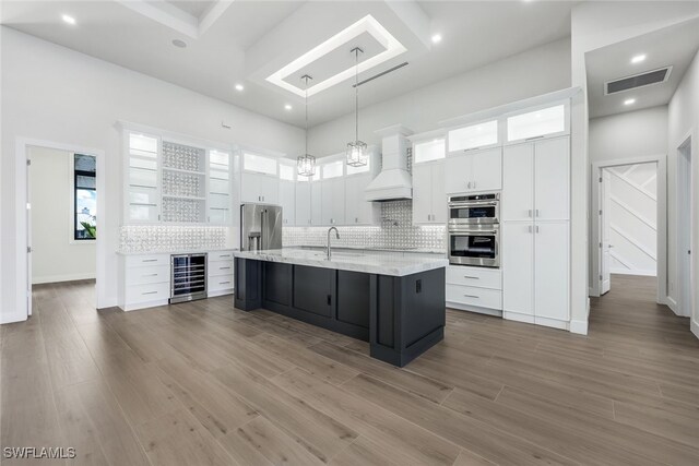 kitchen featuring a kitchen island with sink, wine cooler, appliances with stainless steel finishes, white cabinets, and premium range hood