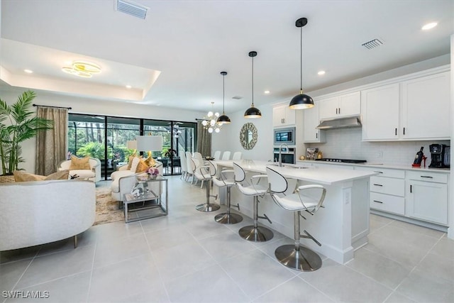 kitchen with pendant lighting, stainless steel microwave, an island with sink, white cabinets, and a raised ceiling