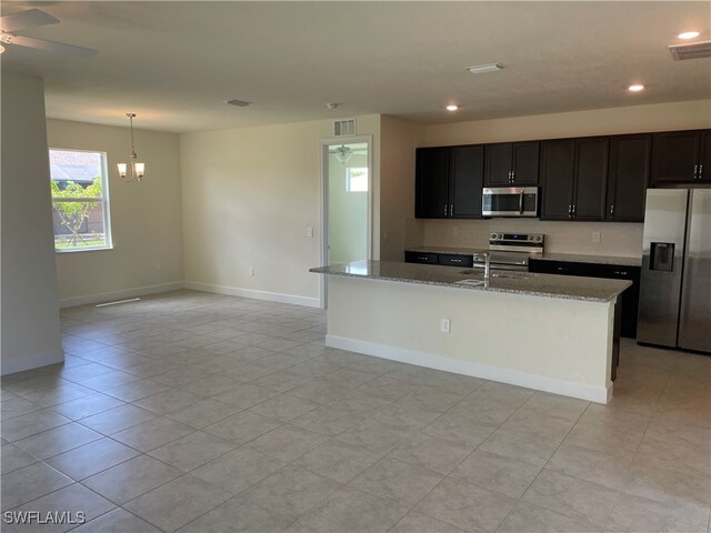 kitchen featuring appliances with stainless steel finishes, a kitchen island with sink, ceiling fan with notable chandelier, pendant lighting, and light stone counters