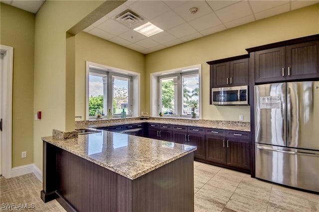 kitchen with light stone countertops, appliances with stainless steel finishes, kitchen peninsula, a drop ceiling, and dark brown cabinets