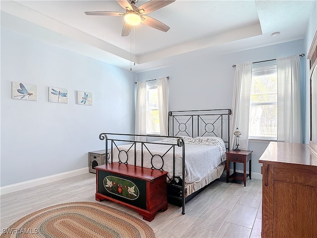 bedroom with ceiling fan, a raised ceiling, multiple windows, and light wood-type flooring