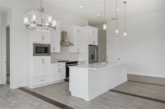 kitchen featuring stainless steel appliances, wall chimney exhaust hood, decorative light fixtures, and a kitchen island with sink