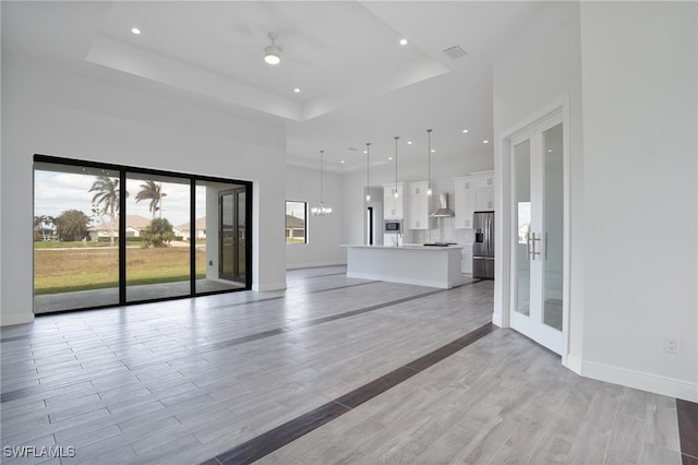 unfurnished living room featuring a high ceiling, ceiling fan with notable chandelier, light wood-type flooring, and a raised ceiling