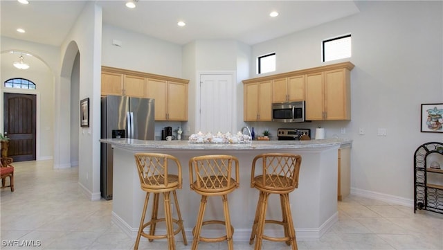 kitchen featuring light brown cabinets, a high ceiling, light stone counters, an island with sink, and appliances with stainless steel finishes