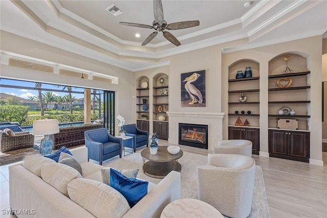 living area featuring a tray ceiling, built in shelves, visible vents, a ceiling fan, and a glass covered fireplace