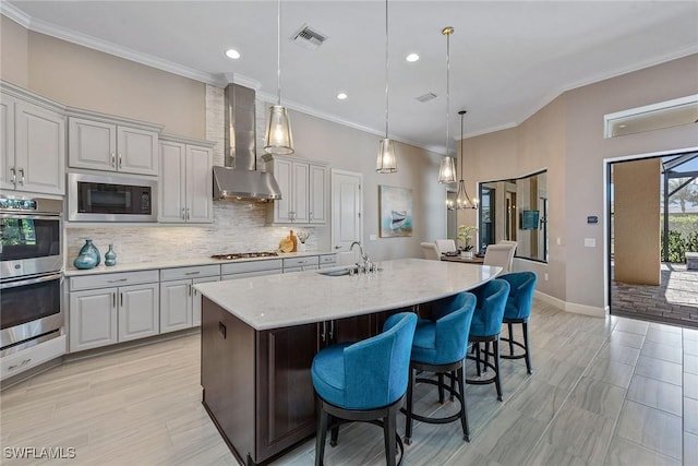 kitchen featuring a kitchen island with sink, stainless steel appliances, tasteful backsplash, a sink, and wall chimney range hood