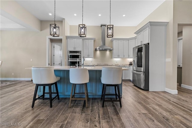 kitchen featuring wall chimney range hood, decorative light fixtures, appliances with stainless steel finishes, and an island with sink