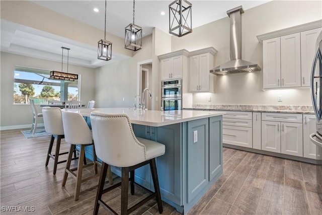 kitchen featuring dark wood-type flooring, wall chimney range hood, double oven, an island with sink, and hanging light fixtures