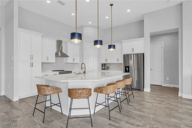 kitchen with wall chimney exhaust hood, a center island with sink, white cabinets, decorative light fixtures, and appliances with stainless steel finishes