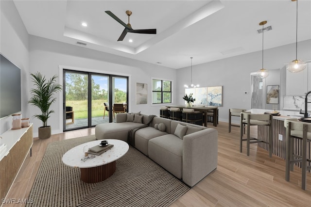 living room with ceiling fan with notable chandelier, a tray ceiling, and light wood-type flooring