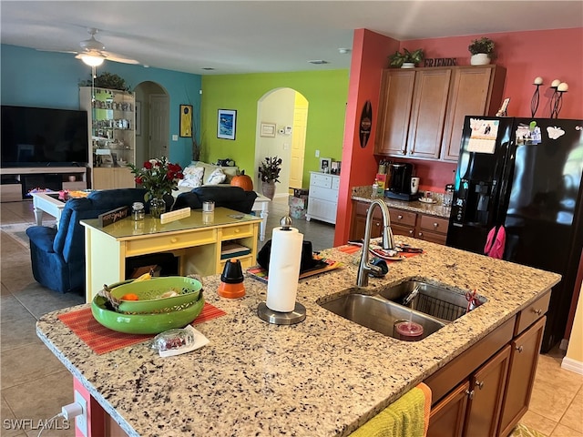 kitchen with sink, ceiling fan, black fridge with ice dispenser, light stone counters, and a kitchen island with sink