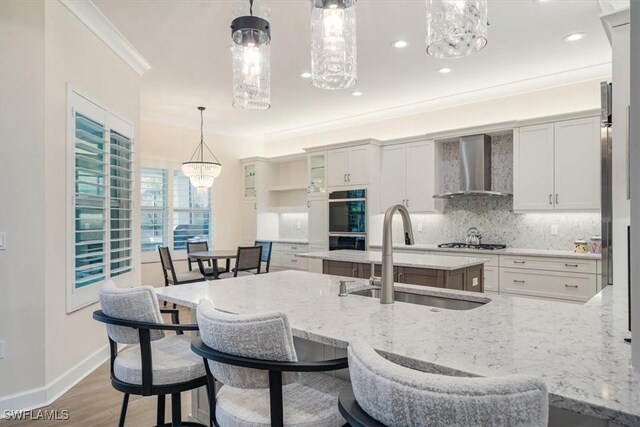 kitchen with wall chimney range hood, tasteful backsplash, a sink, ornamental molding, and a notable chandelier
