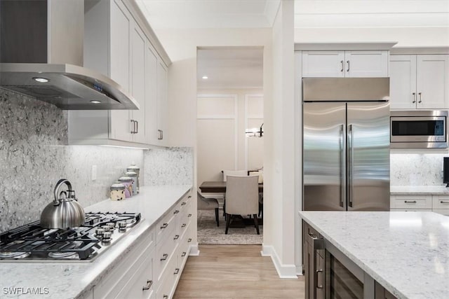 kitchen featuring light stone countertops, light wood-style flooring, decorative backsplash, built in appliances, and wall chimney exhaust hood