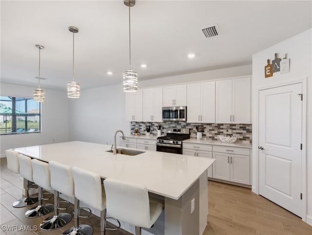 kitchen with appliances with stainless steel finishes, a kitchen island with sink, sink, white cabinetry, and hanging light fixtures