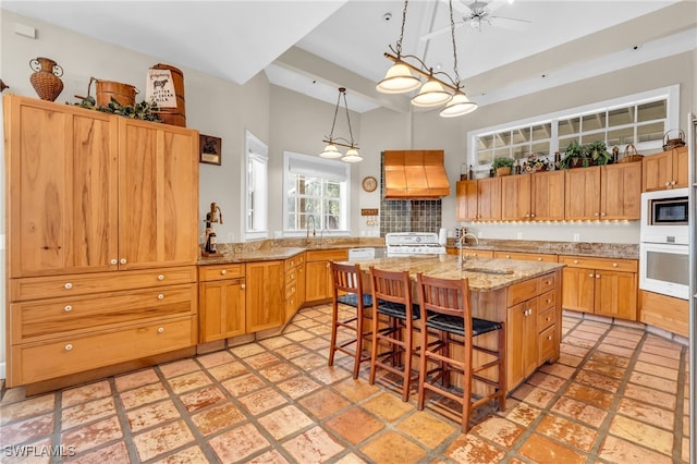 kitchen with a kitchen island with sink, pendant lighting, custom exhaust hood, light stone counters, and white appliances