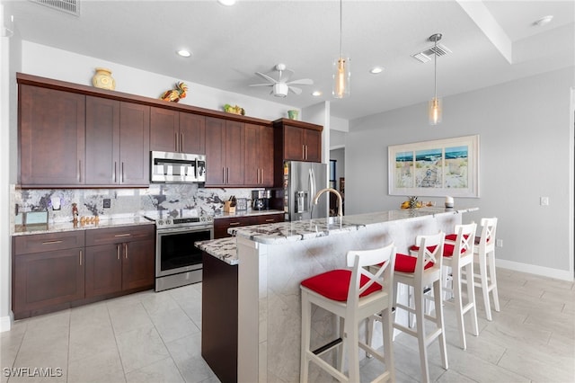 kitchen featuring a kitchen island with sink, stainless steel appliances, backsplash, pendant lighting, and light stone counters