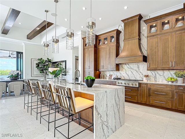 kitchen with pendant lighting, custom exhaust hood, a kitchen island with sink, stainless steel gas cooktop, and light stone counters